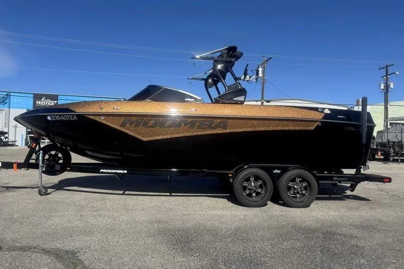 The Image of 2021 Moomba Makai boat on trailer, black and gold, parked outdoors under clear blue sky. - 1