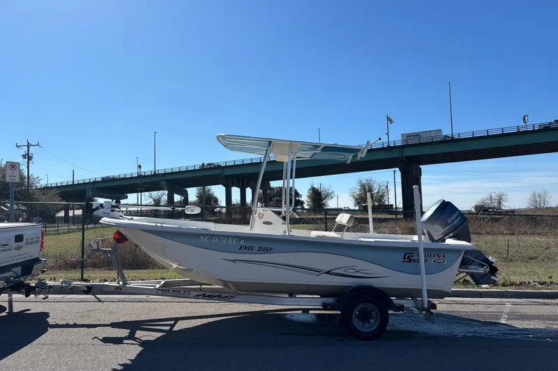 Slide: The Image of 2016 Carolina Skiff 198 DLV boat on trailer, parked near a bridge under clear blue sky. - 2
