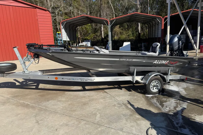 Slide: The Image of 2018 Alumacraft Crappie Deluxe boat on trailer, parked near red storage sheds. - 1