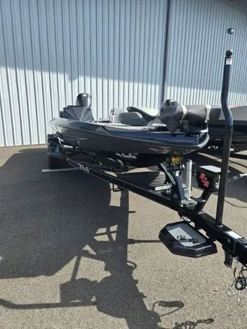 Slide: The Image of 2025 Bass Cat Lynx STS boat on trailer, parked outdoors near a metal building. - 1