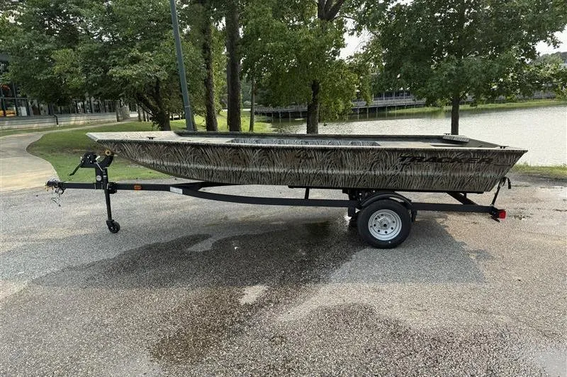 The Image of 2026 Tracker Grizzly 1648 Utility boat on trailer, parked near a lake with trees. - 0