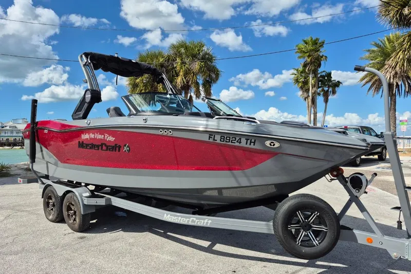 The Image of 2022 MasterCraft X24 boat on trailer, red and gray, parked near palm trees under blue sky. - 1