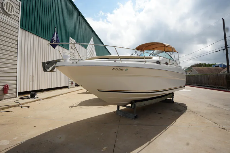 The Image of 2000 Sea Ray 270 Sundancer boat on dry dock, with a green and white building backdrop. - 1