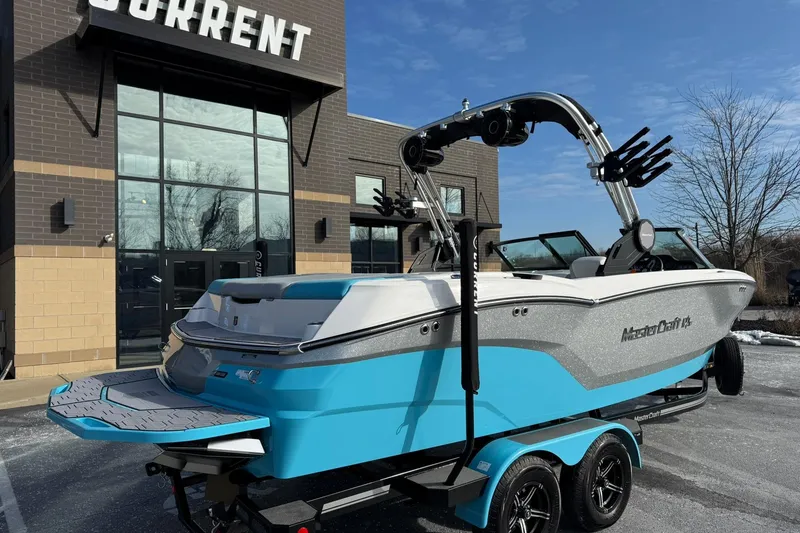 Slide: The Image of 2023 MasterCraft NXT23 boat in blue and white, parked outside a modern building. - 2
