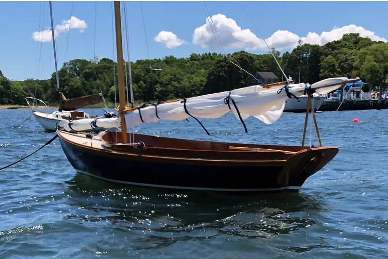 Slide: The Image of 2018 Ballentine's Boat Shop Doughdish sailboat on calm water, with trees in the background. - 4