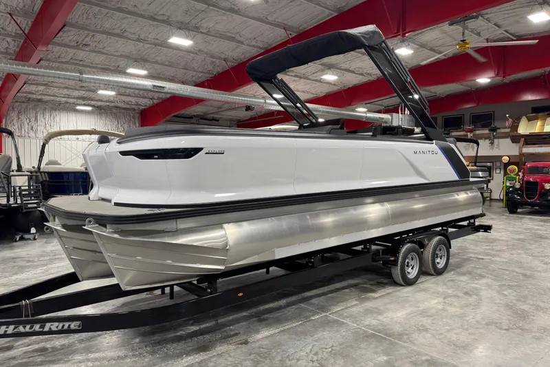 The Image of 2025 Manitou 24 Explorer pontoon boat on trailer in indoor showroom. - 1