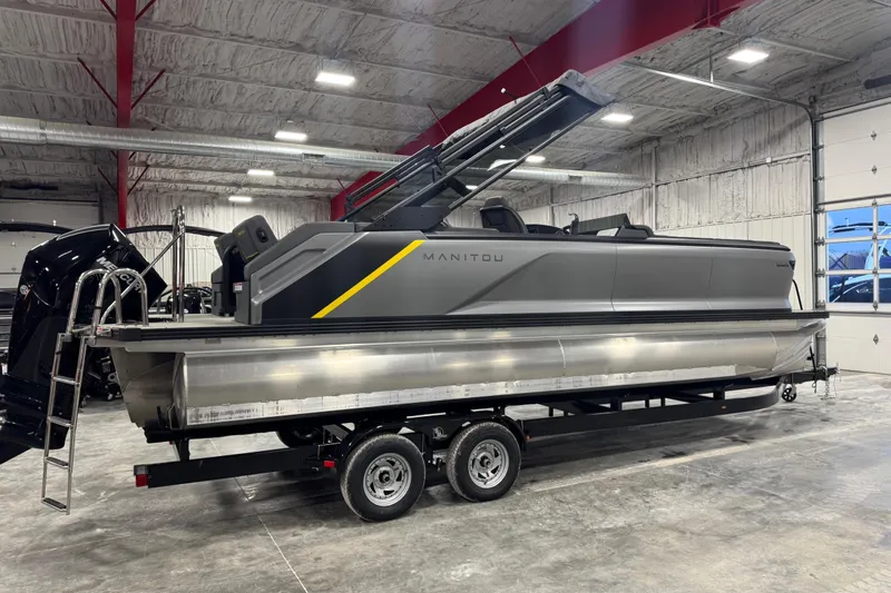 Slide: The Image of 2024 Manitou 24 Explorer pontoon boat on trailer in a spacious indoor facility. - 2