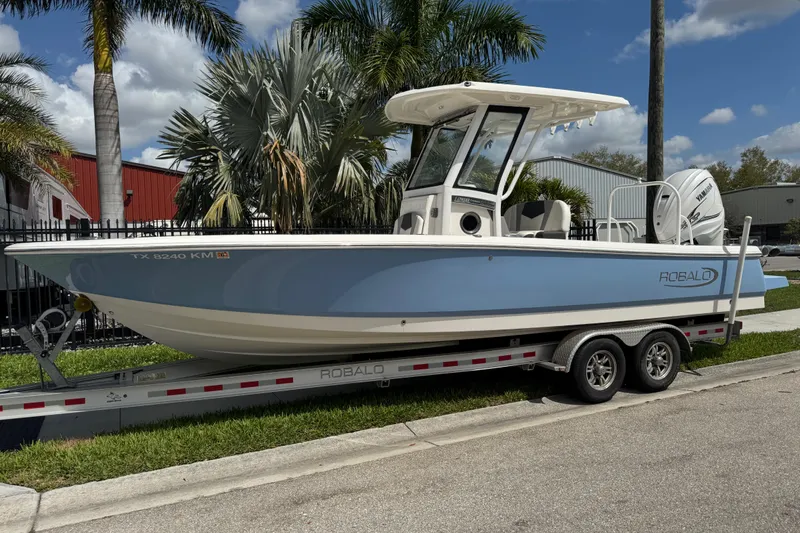 The Image of 2022 Robalo 266 Cayman boat on trailer, parked near palm trees and industrial buildings. - 0