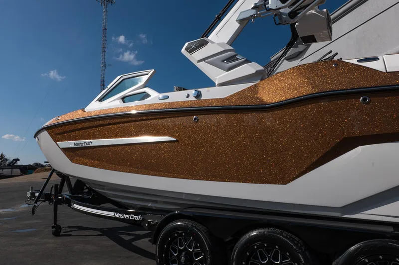 Slide: The Image of 2026 MasterCraft X24 boat with glittering gold exterior on a trailer under blue sky. - 26