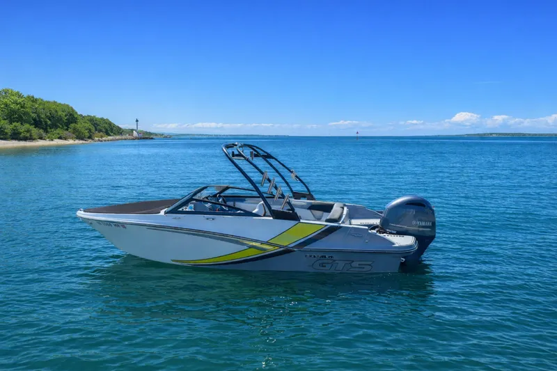 Slide: The Image of 2020 Glastron GTS 200 boat on clear blue water near a lush green shoreline. - 2