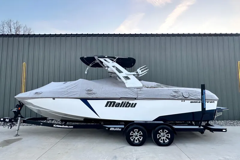 Slide: The Image of 2017 Malibu Wakesetter 25 LSV boat covered on trailer, parked by a metal building. - 20