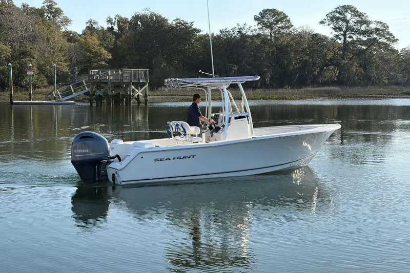 Slide: The Image of 2014 Sea Hunt Triton 225 boat cruising on calm water near a wooded shoreline. - 29