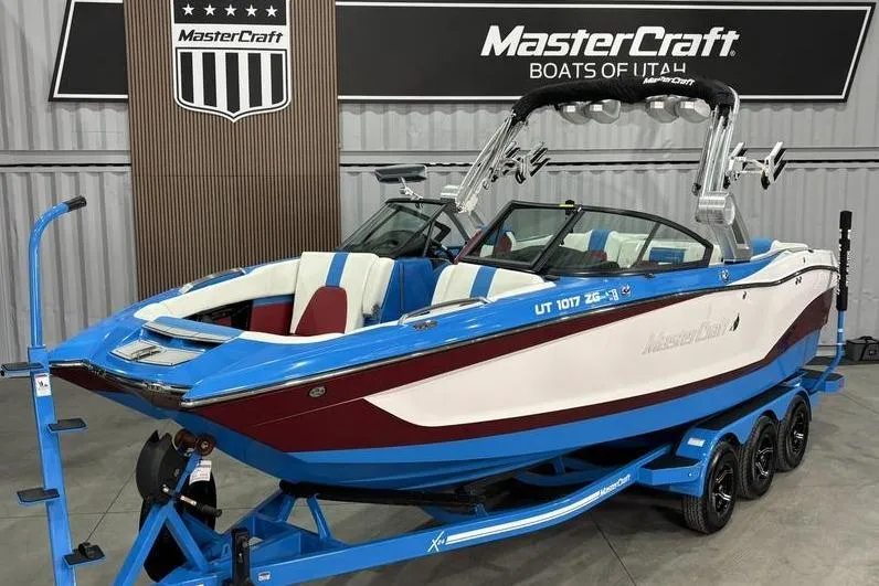 The Image of 2024 MasterCraft X24 boat in showroom, blue and white design, on trailer. - 1