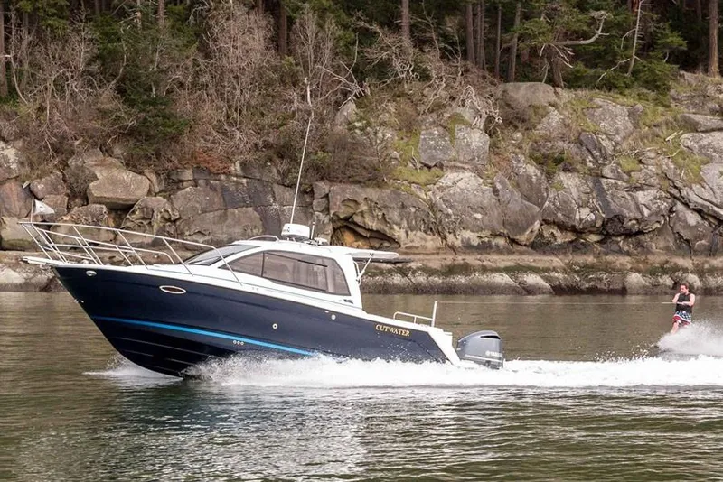 Slide: The Image of Manufacturer Provided Image: 2022 Cutwater C-24 Coupe cruising near rocky shoreline with a person wakeboarding. - 2