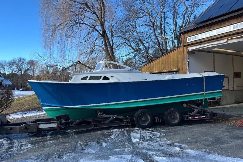 Slide: The Image of 1970 Brownell Cuttyhunk boat on trailer, blue and green hull, parked near a wooden building. - 7