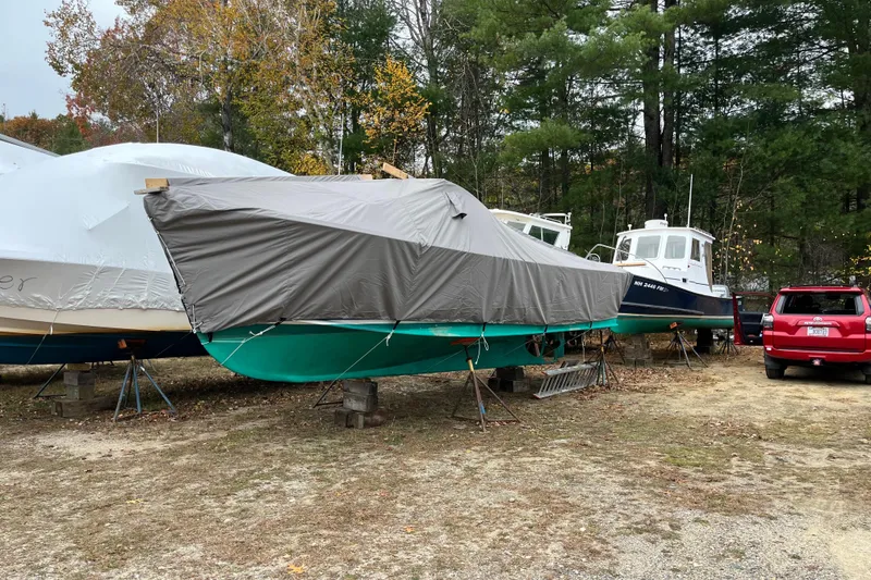 Slide: The Image of 1970 Brownell Cuttyhunk boat covered and stored outdoors, surrounded by trees and vehicles. - 8