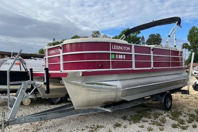 The Image of 2022 Lexington HP320 pontoon boat on trailer, red and silver design, parked outdoors. - 0