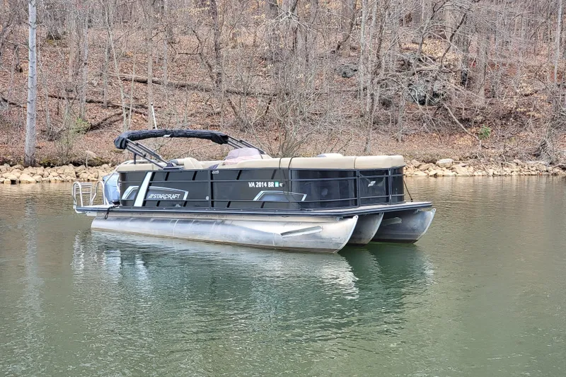 Slide: The Image of 2024 Starcraft EXS 3 pontoon boat on a calm lake with wooded shoreline. - 6