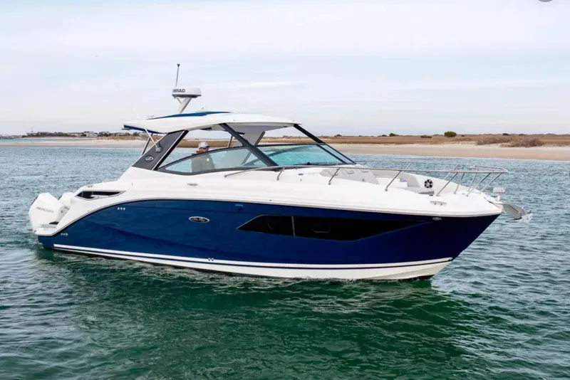 The Image of 2024 Sea Ray Sundancer 320 Outboard cruising on calm waters near a sandy shoreline. - 0