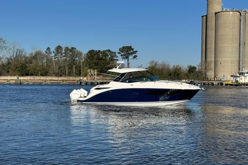 Slide: The Image of 2024 Sea Ray Sundancer 320 Outboard cruising on a calm river near industrial silos. - 6
