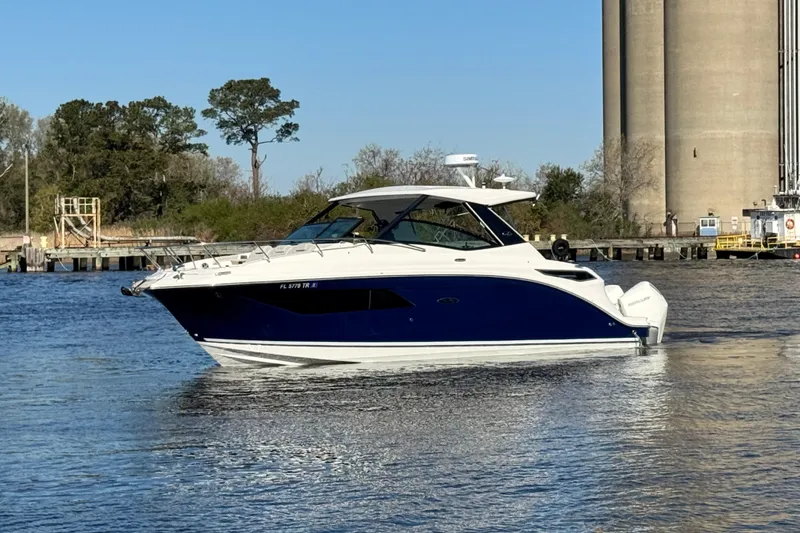 Slide: The Image of 2024 Sea Ray Sundancer 320 Outboard cruising on a calm river near industrial structures. - 7