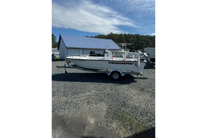 The Image of 2009 Triumph 1700 Skiff boat on trailer, parked near a blue-roofed building. - 0