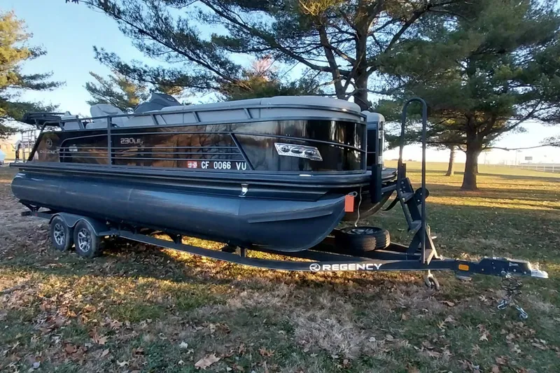 The Image of 2021 Regency 230 LE3 Sport pontoon boat on trailer, parked in a grassy area. - 0