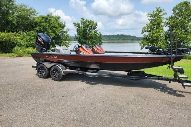 The Image of 2024 Avid 21 XB Elite boat on trailer by a lake, under a partly cloudy sky. - 0