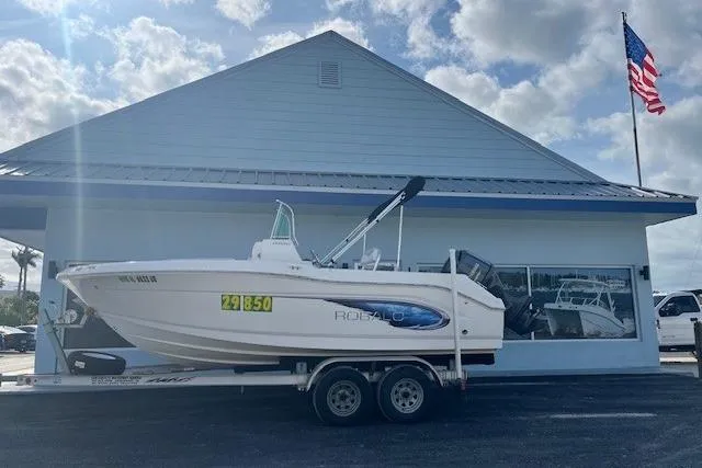 The Image of 2014 Robalo R180 Center Console boat on trailer, parked outside a building with American flag. - 1