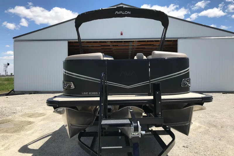 Slide: The Image of 2026 Avalon LSZ Quad Lounge Tri Log pontoon boat on trailer, parked outside a white building. - 10