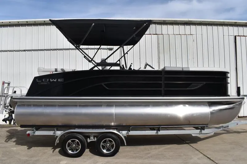 The Image of 2026 Lowe SS 210 pontoon boat on trailer, black canopy, parked by a white building. - 1