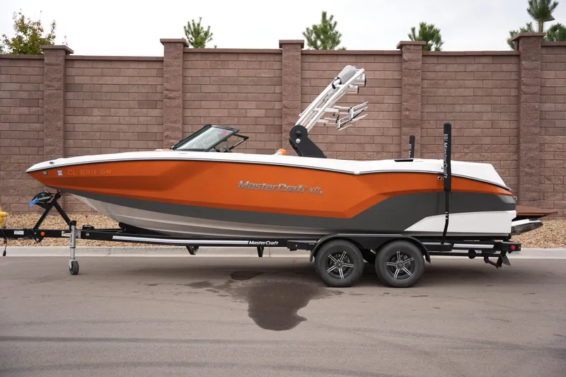 The Image of 2021 MasterCraft NXT24 boat on trailer, orange and gray design, parked by brick wall. - 1