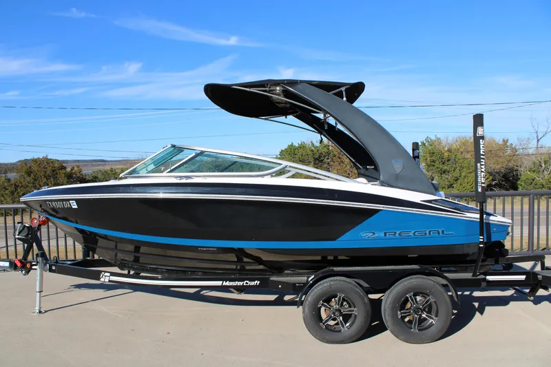 The Image of 2017 Regal 2100 Bowrider boat on trailer, black and blue design, clear sky background. - 1