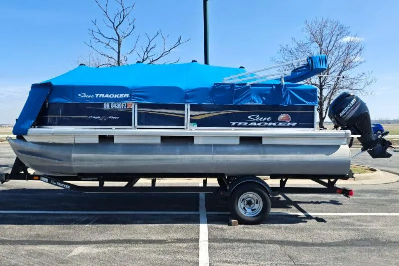 Slide: The Image of 2020 SUNTRACKER pb18 pontoon boat with blue cover on trailer in parking lot. - 17