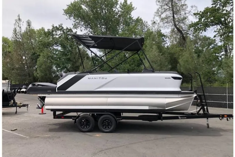 Slide: The Image of 2025 Manitou Cruise pontoon boat on trailer, parked outdoors with trees in background. - 0