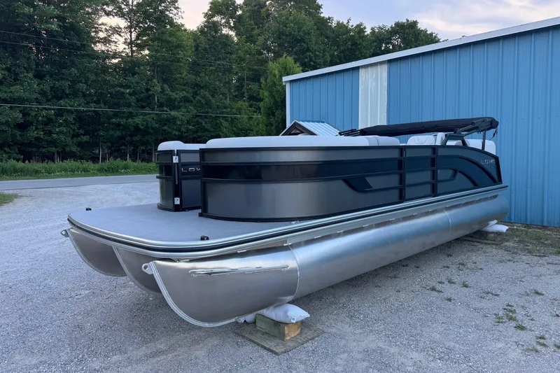 The Image of 2025 Lowe SS230WV pontoon boat parked near a blue building, surrounded by trees. - 0