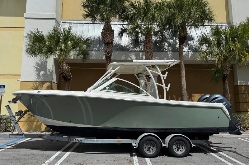 Slide: The Image of 2018 Sailfish 275 DC boat on trailer, parked near palm trees. - 2