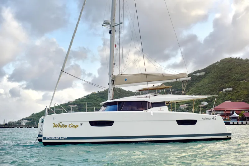 The Image of 2024 Fountaine Pajot Astrea 42 catamaran sailing near lush green hills and cloudy sky. - 0