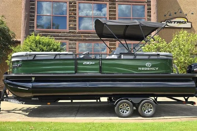 The Image of 2026 Regency 230 DL pontoon boat on trailer, green and black design, parked outdoors. - 0