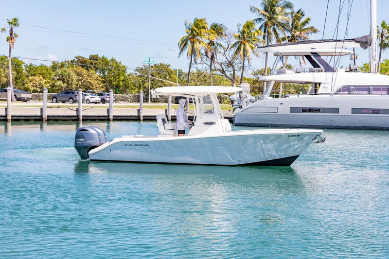 Slide: The Image of 2023 Cobia 240 Center Console boat cruising in a sunny marina with palm trees. - 2