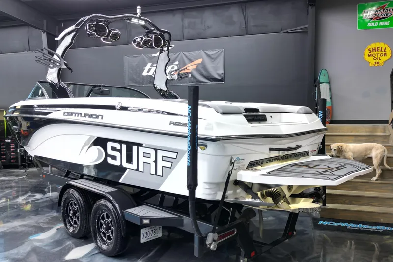 Slide: The Image of 2017 Centurion Ri237 surf boat on trailer in showroom with surfboard and dog nearby. - 25