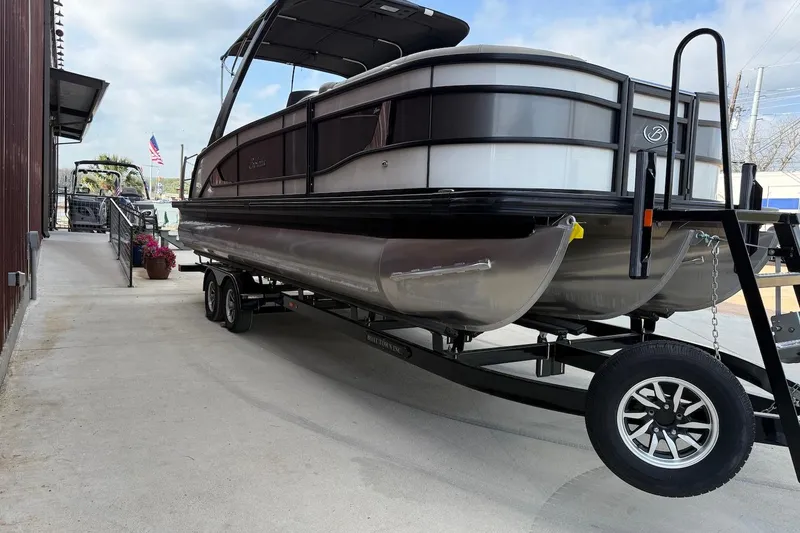 Slide: The Image of 2022 Barletta LUSSO 25UEA pontoon boat on trailer, parked near a building. - 2