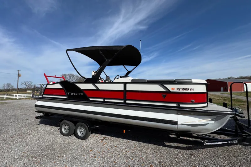 Slide: The Image of 2024 Trifecta 25ULE SS pontoon boat on trailer, red and white design, parked outdoors. - 43