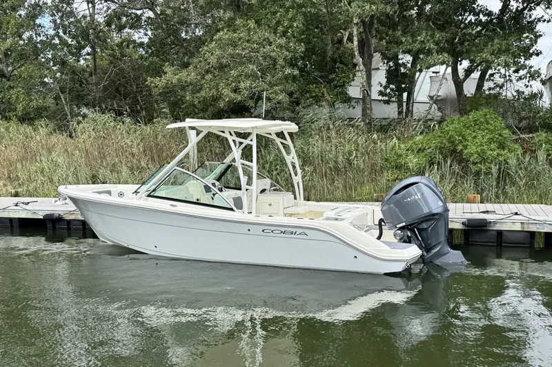 Slide: The Image of 2026 Cobia 240 Dual Console boat docked by lush greenery. - 2