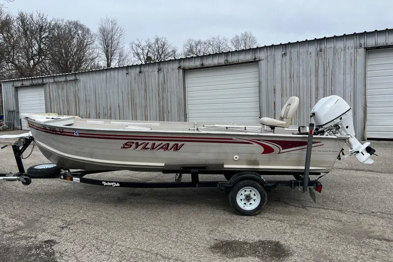 The Image of 2004 Sylvan 1600 Super Snapper boat on trailer, parked near metal garages. - 1