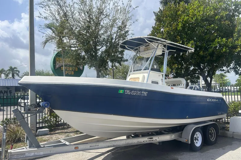 The Image of 2009 Cobia 216 Center Console boat on trailer, parked outdoors near trees and a sports pub sign. - 0