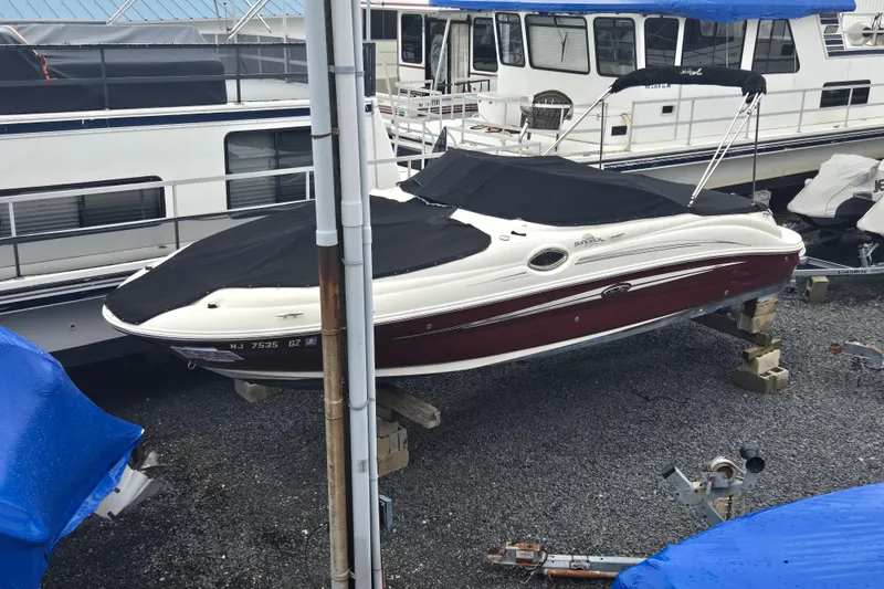 The Image of 2007 Sea Ray 240 Sundeck boat with black cover, docked on gravel. - 0