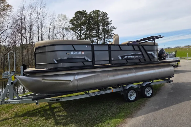 Slide: The Image of 2024 Starcraft EXS 1 pontoon boat on trailer, parked near a lake. - 1