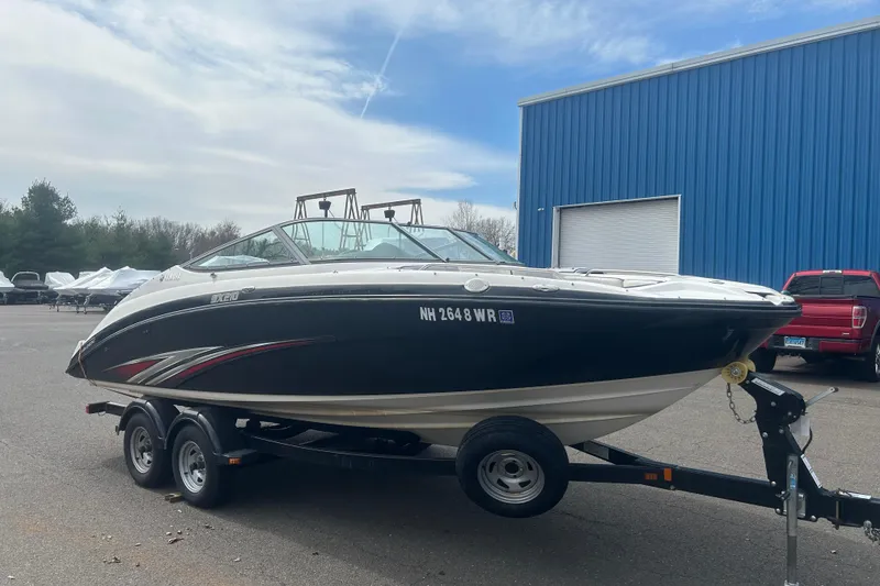 Slide: The Image of 2015 Yamaha Boats SX210 on trailer, parked near blue building under cloudy sky. - 3