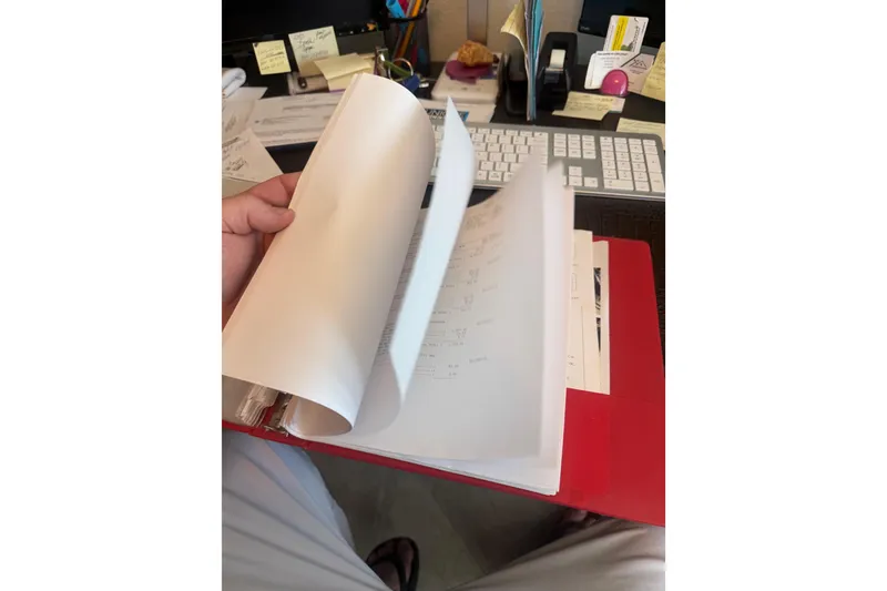 Slide: The Image of Person flipping through documents in a red binder on a cluttered desk. - 76